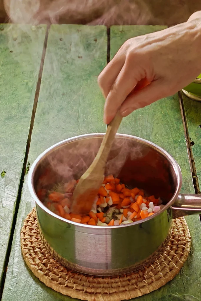 A raffia trivet with a stainless steel pot lies on a green wooden table. In it is the ready-cooked mixture for the Chestnut Spread. Hot steam rises from the pot.