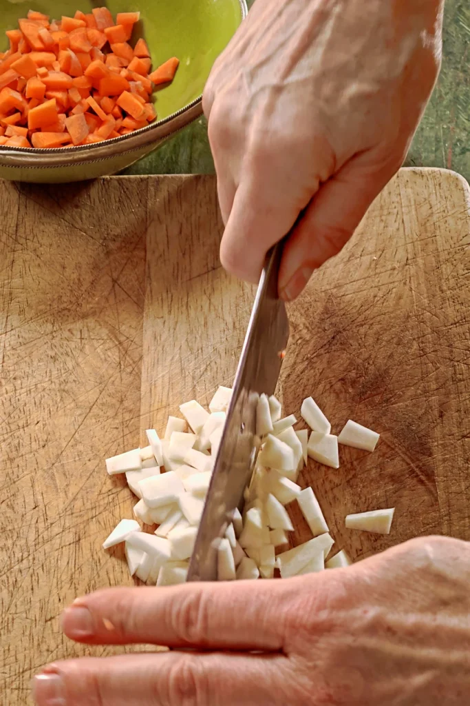 Chopped celery lies on a wooden board, two hands hold a large knife and cut the celery into small pieces. In the background is a green bowl with chopped carrots.