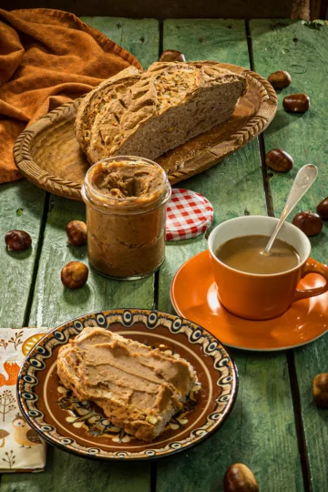 The snack is served on a green wooden table: a loaf of bread with créme de marrons. The bread lies on a brown plate. Behind is cup of coffee and a preserving jar with the créme de marrons. Further in the background is a flat wooden bowl with a sliced loaf of bread and a orange linen kitchen towel. Scattered around everything are fresh chestnuts in shells.