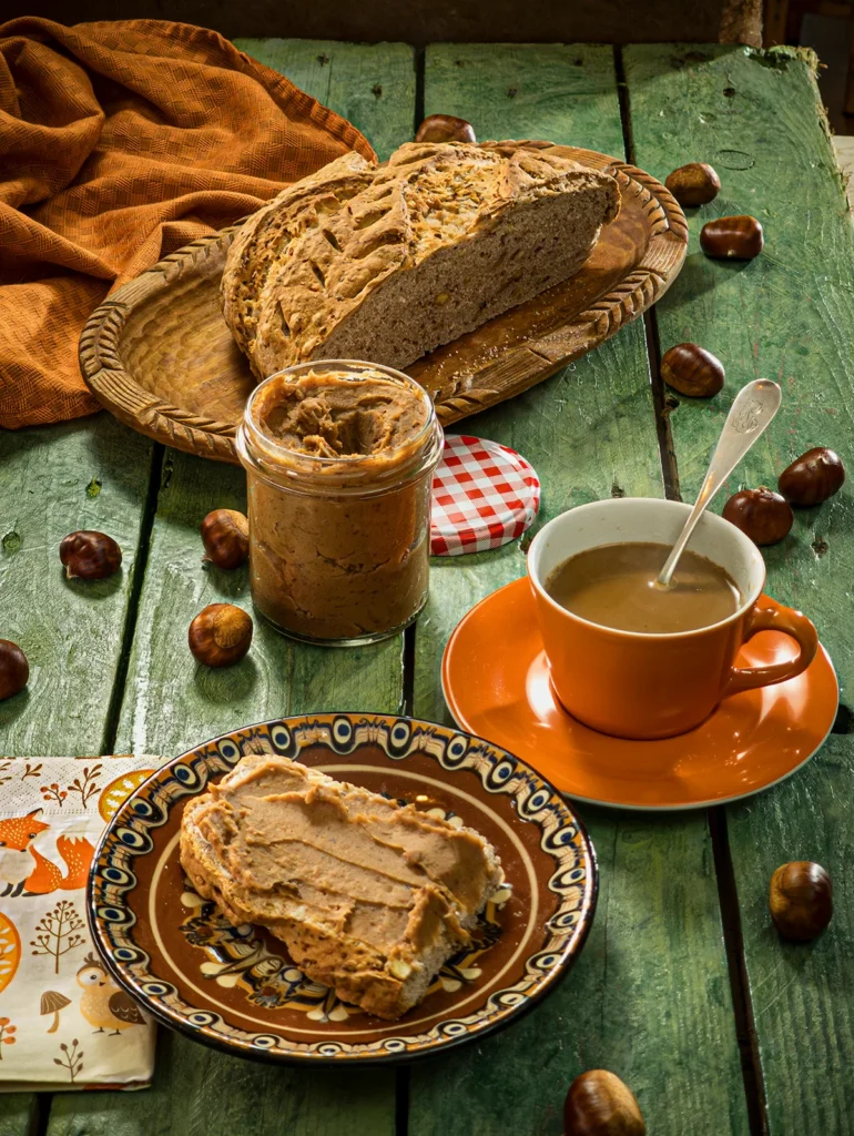 The snack is served on a green wooden table: a loaf of bread with créme de marrons. The bread lies on a brown plate. Behind is cup of coffee and a preserving jar with the créme de marrons. Further in the background is a flat wooden bowl with a sliced loaf of bread and a orange linen kitchen towel. Scattered around everything are fresh chestnuts in shells.