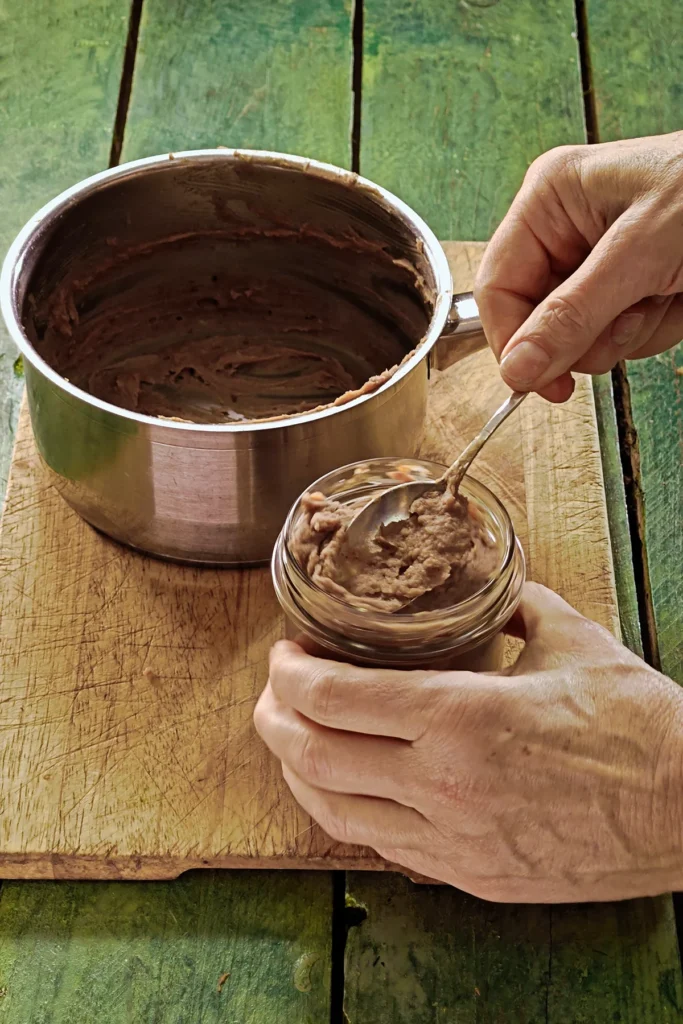 A wooden cutting board lies on a green wooden table, on it stands a stainless steel pot . The pot contains the pureed crème de marrons. In the foreground, one hand holds a jar filled with crème de marrons. The other hand holds a spoon and presses the cream into the jar.