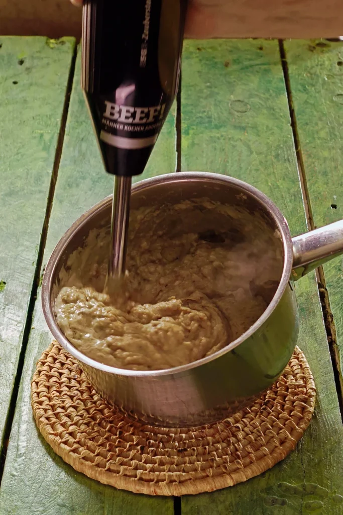 A raffia trivet with a stainless steel pot lies on a green wooden table. In it are the blended ingredients for the Chestnut Spread. A hand blender purees the mixture into a fine cream.