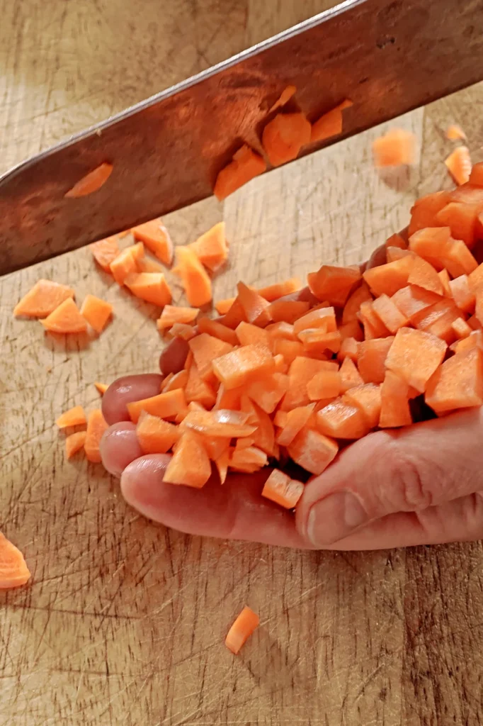 The palm of one hand is filled with a mountain of finely chopped carrots. The hand lies on a wooden board, the blade of a large knife hovers in the air.