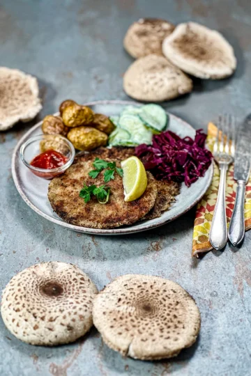 A beautiful grey plate with two parasol schitzel, braised red cabbage, cucumber salad, and potatoes. Fresh parasol mushrooms are presented around the plate.