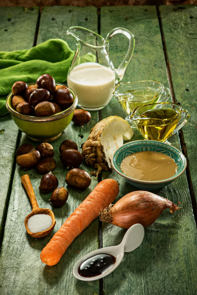 On a green wooden table are the ingredients for chestnut spread, a green bowl with fresh chestnuts, a glass jug with soy cream, two smaller jugs with rapeseed oil and olive oil, a bowl with tahini, a piece of celery, a carrot, a shallot, a small bowl with sugar beet molasses. A roughly carved wooden spoon carries salt. A few chestnuts are scattered among the bowls and vegetables. A green linen kitchen towel lies in the background.