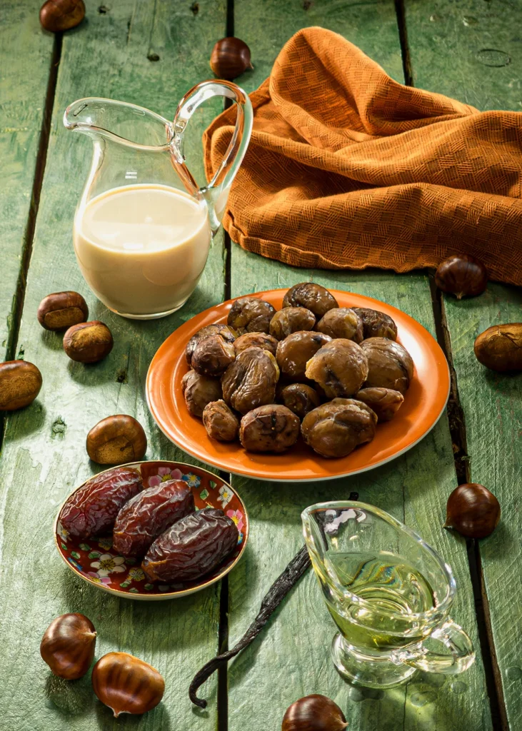 The ingredients for crème de marrons are laid out on a green wooden table. In the foreground is a glass of rapeseed oil, a vanilla pod and a small plate of dates. Behind them are an orange plate with already cooked chestnuts, a glass jug of oat milk and an orange kitchen towel. Fresh chestnuts are scattered around the scene.