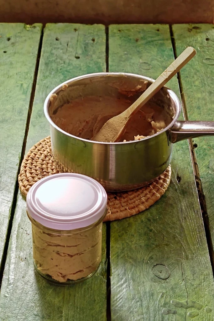 A raffia trivet with a stainless steel pot lies on a green wooden table. In it lies a wooden spatula in the chestnut spread. In front of it is a preserving jar with a white lid. In it is the preserved chestnut spread.