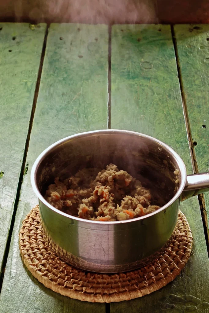 A raffia trivet with a stainless steel pot lies on a green wooden table. In it is the ready-cooked mixture for the Chestnut Spread. Hot steam rises from the pot.