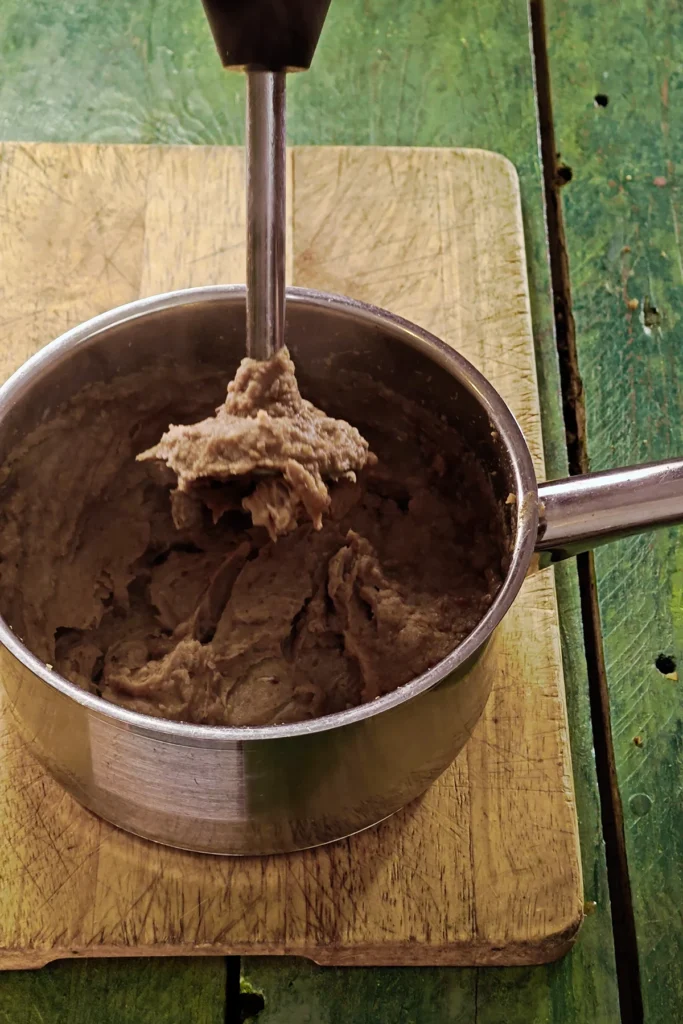 A wooden cutting board lies on a green wooden table with a stainless steel pot on top. The pot contains pureed crème de marrons, a hand blender has just finished blending.