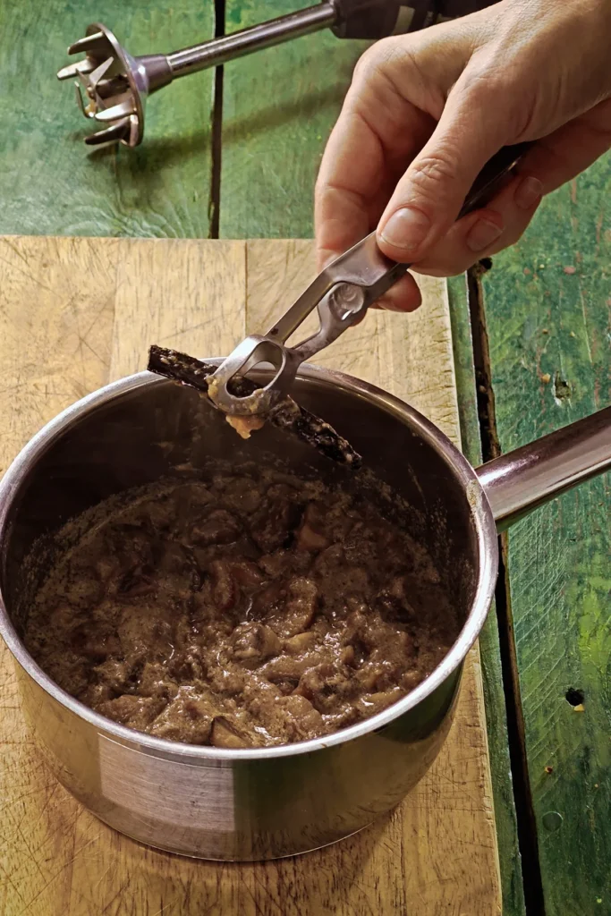 A wooden cutting board lies on a green wooden table with a stainless steel pot on top. A hand with a kitchen tongs removes the vanilla pod from the freshly cooked mixture for créme de marrons.