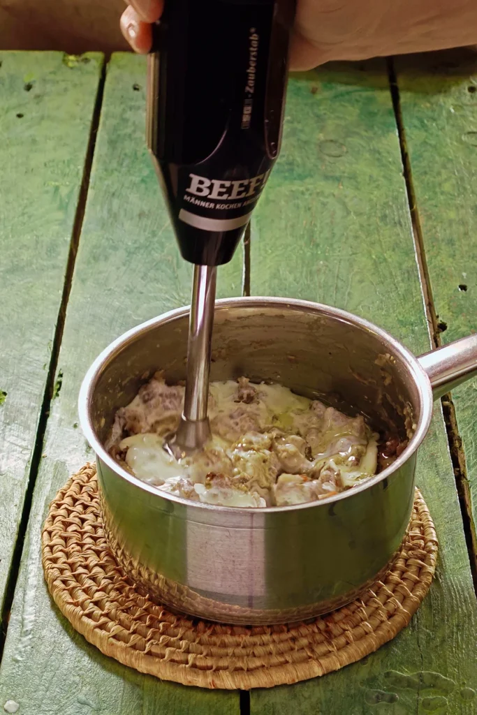 A raffia trivet with a stainless steel pot lies on a green wooden table. In it are the cooked ingredients for the Chestnut Spread. A hand blender purées the mixture into a puree.