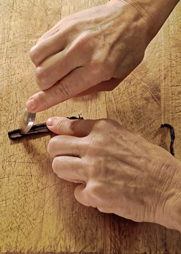 A wooden chopping board lies on a green wooden table with dates. Hands hold the vanilla pod and the pulp is scraped out with a small spoon.