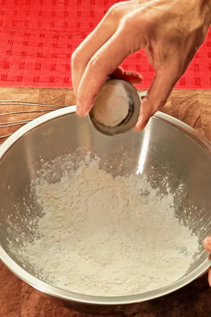 A hand pours baking powder from a little bowl into a stainless steel bowl. The background is a red tablecloth.