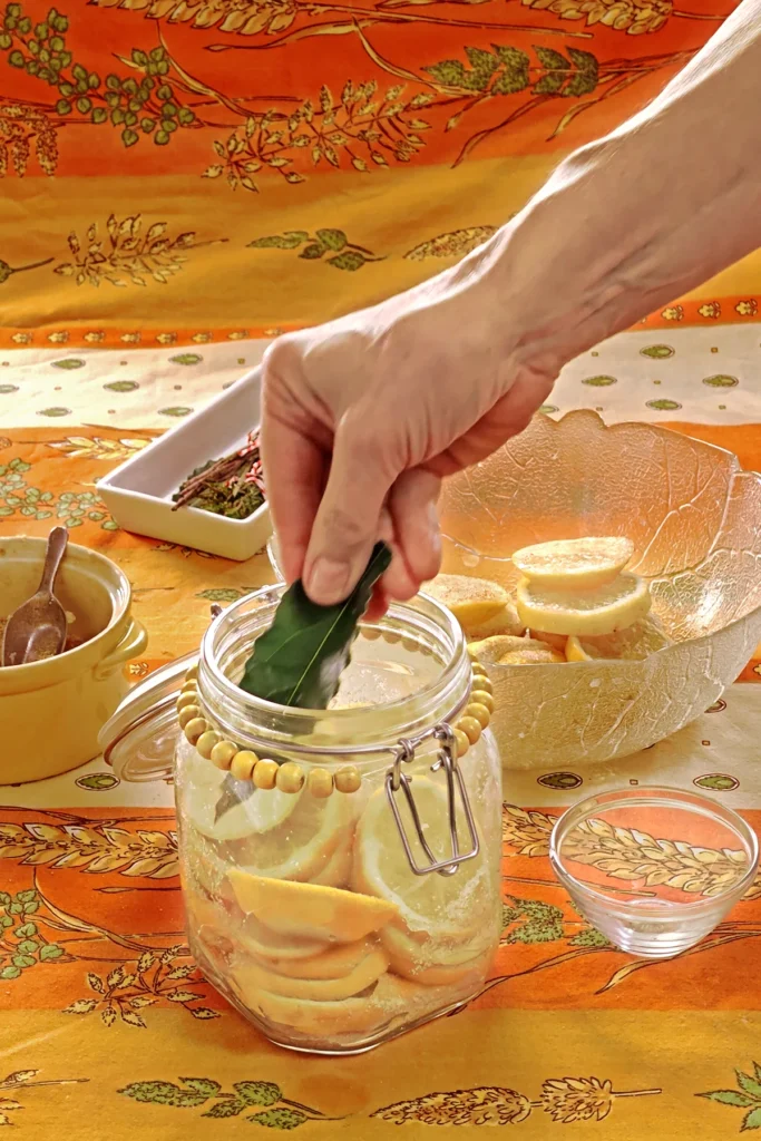 A hand is adding a bay leaf in the preserving jar with lemon slices. Next to it is a small bowl that will be used as a weight to press the lemon slices together. Behind the jar is a glass bowl with lemon slices, a white bowl with herbs, and a yellow bowl with the salt and sugar mixture, all set on an orange tablecloth with wheat and oat straw motifs.