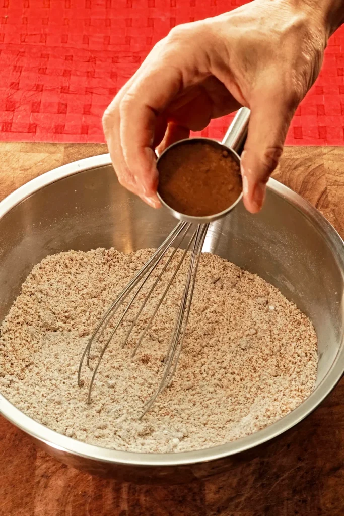 A hand pours Ceylon cinnamon from a little bowl into a stainless steel bowl containing a whisk. The background is a red tablecloth.