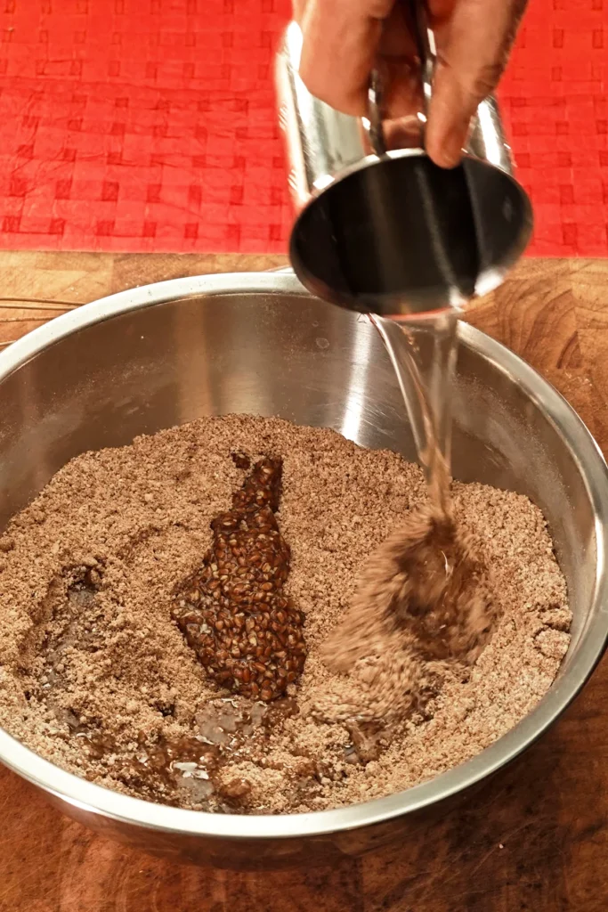 A hand pours coconut oil from a metal jug into a stainless steel bowl. The background is a red tablecloth.