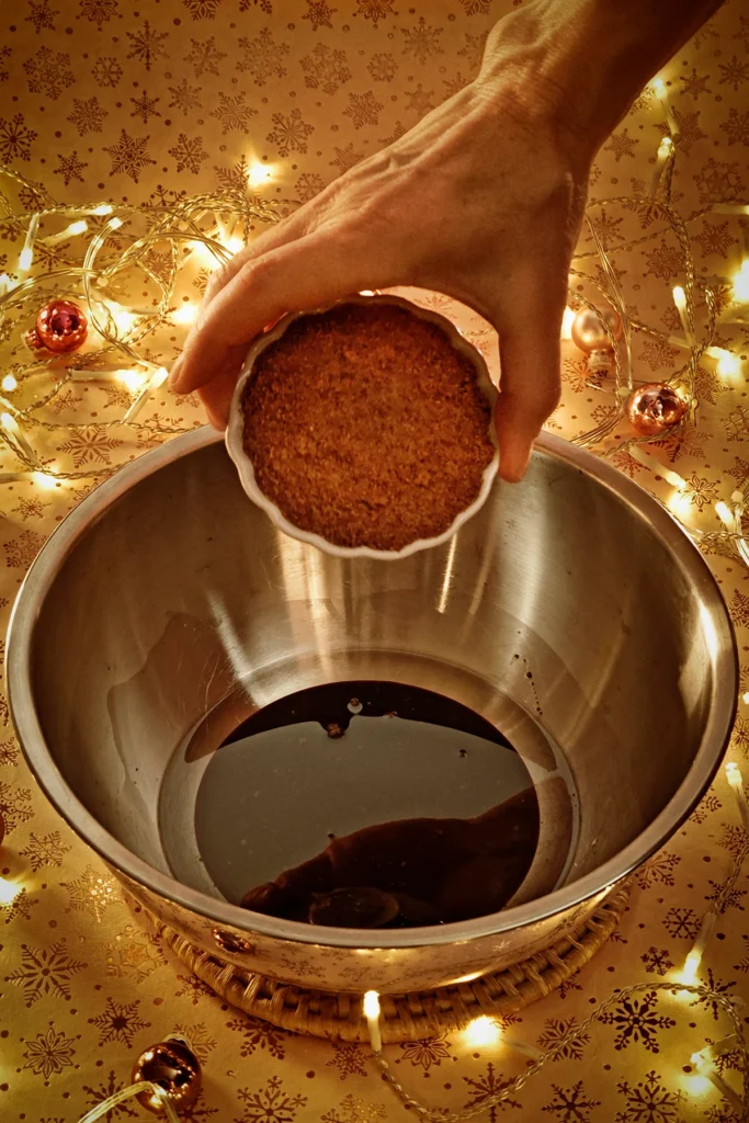 A hand adds coconut sugar from a bowl to the stainless steel bowl. A string of lights shines in the background, and a few small Christmas tree baubles are scattered about. The light background has a festive gold star motif.