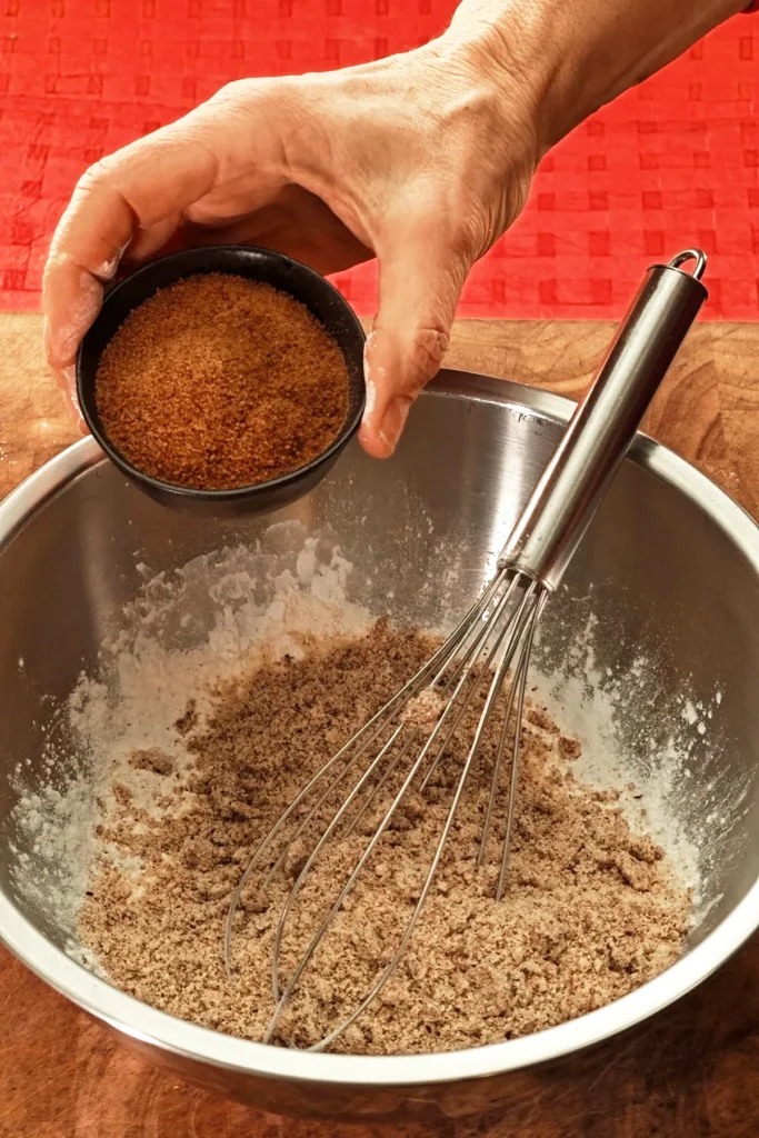 A hand pours coconut sugar from a bowl into a stainless steel bowl containing a whisk. The background is a red tablecloth.