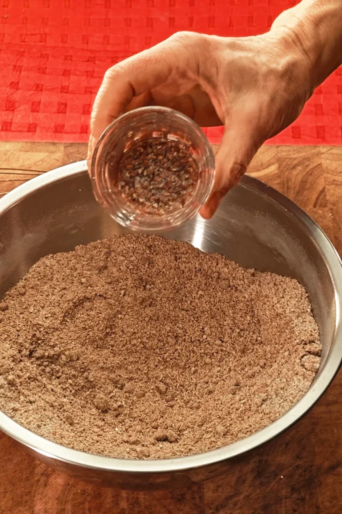 A hand pours swollen flaxseed from a glass into a stainless steel bowl. The background is a red tablecloth.