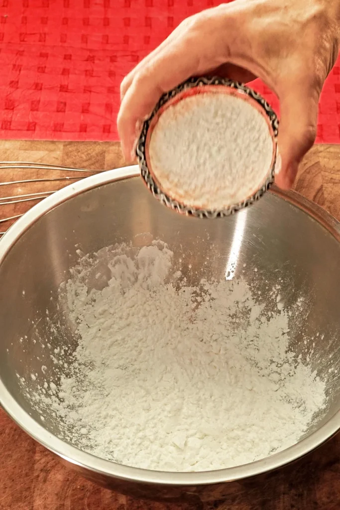 A hand pours flour from a bowl into a stainless steel bowl containing a whisk. The background is a red tablecloth.