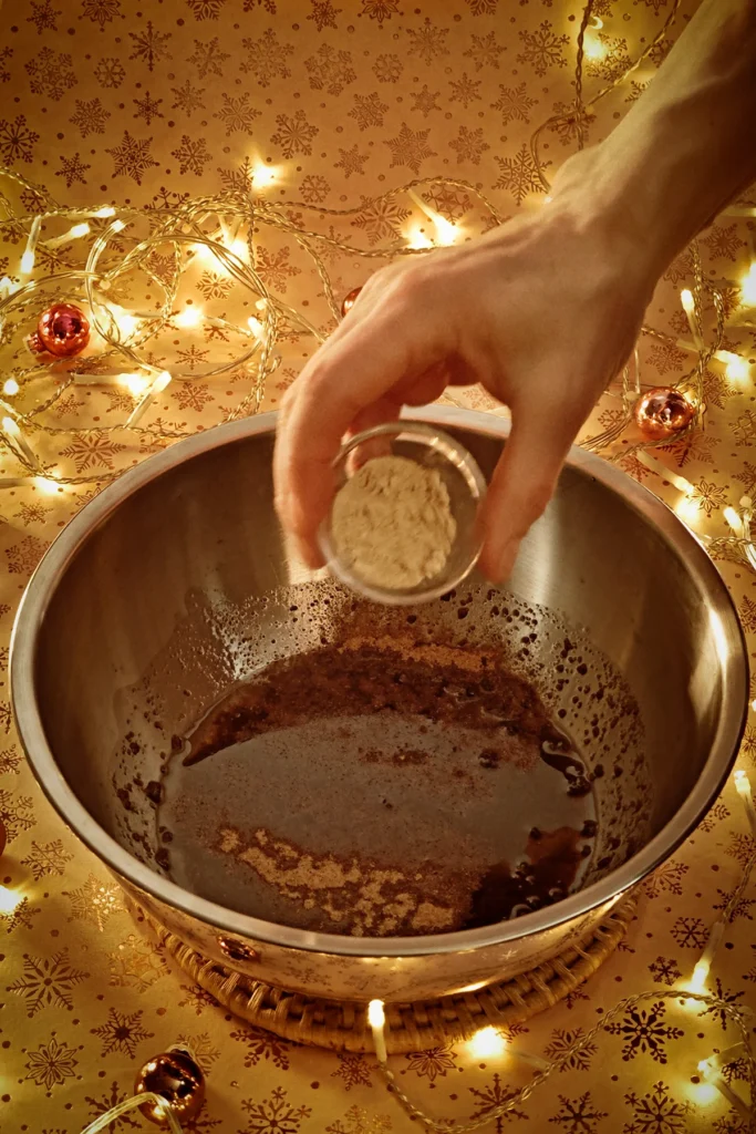 A hand adds ginger powder from a metal bowl to the stainless steel bowl. A string of lights shines in the background, and a few small Christmas tree baubles are scattered about. The light background has a festive gold star motif.