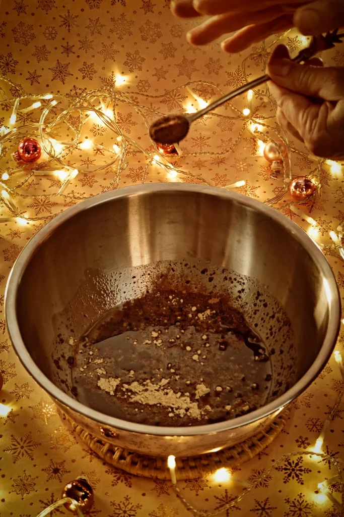 A hand adds cinnamon to the stainless steel bowl with a spoon. A string of lights shines in the background, and a few small Christmas tree baubles are scattered about. The light background has a festive gold star motif.