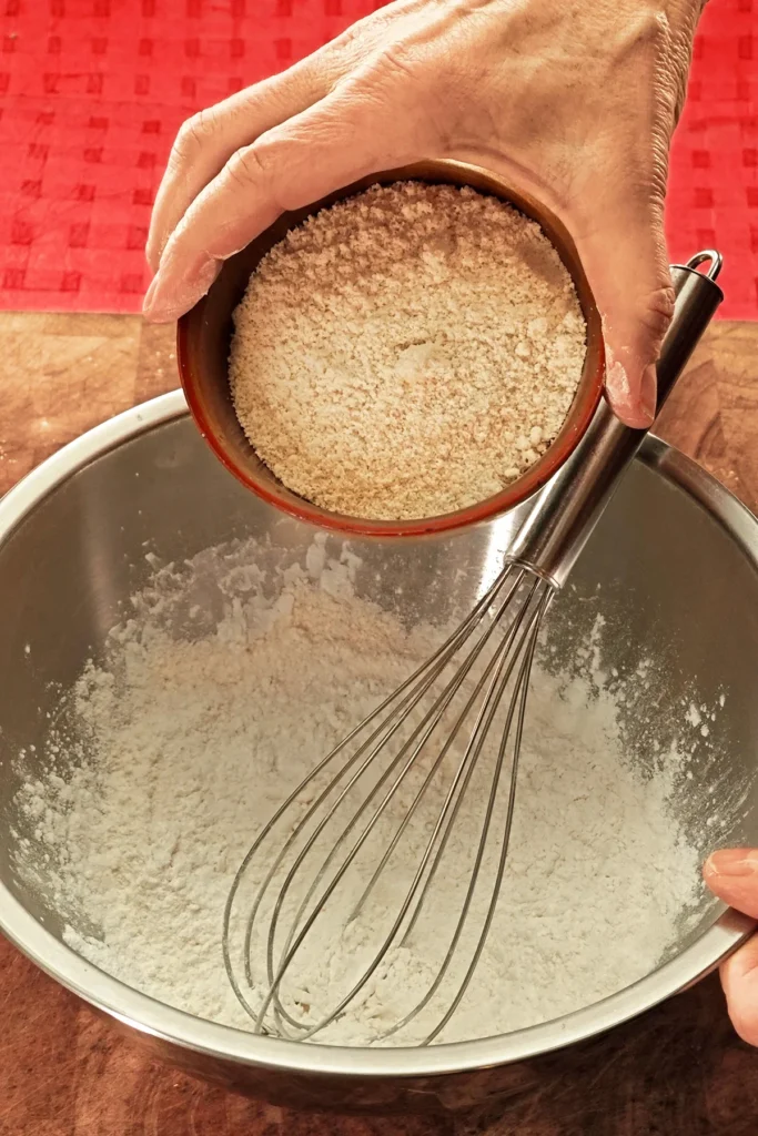 A hand pours ground almonds from a bowl into a stainless steel bowl containing a whisk. The background is a red tablecloth.