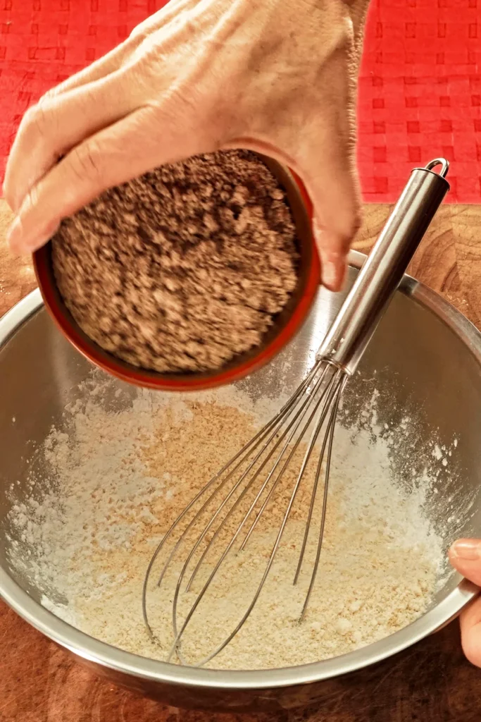 A hand pours ground hazelnuts from a bowl into a stainless steel bowl containing a whisk. The background is a red tablecloth.