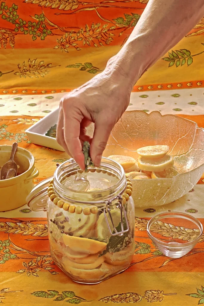 A hand is filling herbs in the preserving jar with lemon slices. Next to it is a small bowl that will be used as a weight to press the lemon slices together. Behind the jar is a glass bowl with lemon slices, a white bowl with herbs, and a yellow bowl with the salt and sugar mixture, all set on an orange tablecloth with wheat and oat straw motifs.