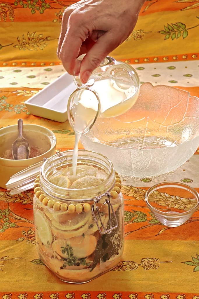 A hand is filling the preserving jar with lemon juice from a glass jug. Next to it is a small bowl used as a weight to press the lemon slices together. Behind the jar is a glass bowl with lemon slices, a white bowl with herbs, and a yellow bowl with the salt and sugar mixture, all set on an orange tablecloth with wheat and oat straw motifs.