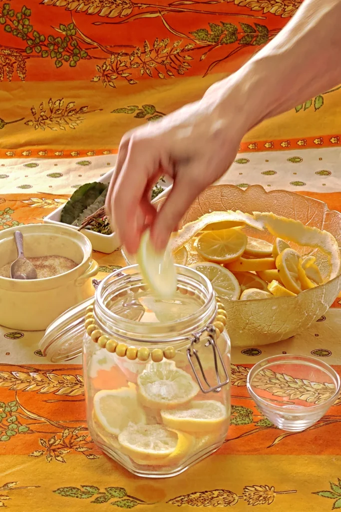 A hand is filling the preserving jar with lemon slices. Next to it is a small bowl used as a weight to press the lemon slices together. Behind the jar is a glass bowl with lemon slices, a white bowl with herbs, and a yellow bowl with the salt and sugar mixture, all set on an orange tablecloth with wheat and oat straw motifs.