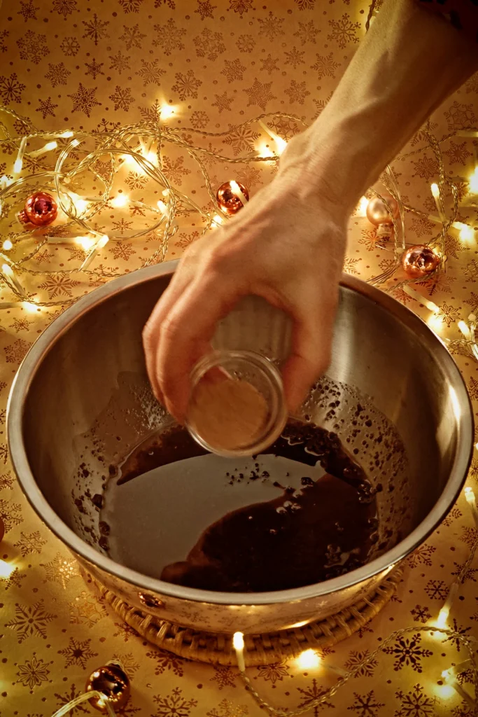 A hand adds ground nutmeg from a glass bowl to the stainless steel bowl. A string of lights shines in the background, and a few small Christmas tree baubles are scattered about. The light background has a festive gold star motif.