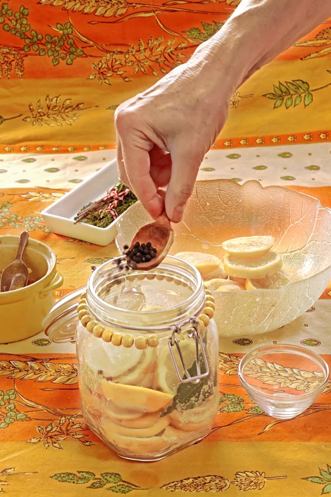 The preserving jar is filled with lemon slices and herbs. One hand holds a wooden spoon and adds peppercorns to the jar. Next to it is a small bowl used as a weight to press the lemon slices together. Behind the jar is a glass bowl with lemon slices, a white bowl with herbs, and a yellow bowl with the salt and sugar mixture, all set on an orange tablecloth with wheat and oat straw motifs.