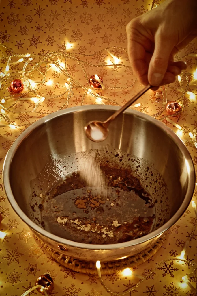 A hand adds salt to the stainless steel bowl with a spoon. A string of lights shines in the background, and a few small Christmas tree baubles are scattered about. The light background has a festive gold star motif.