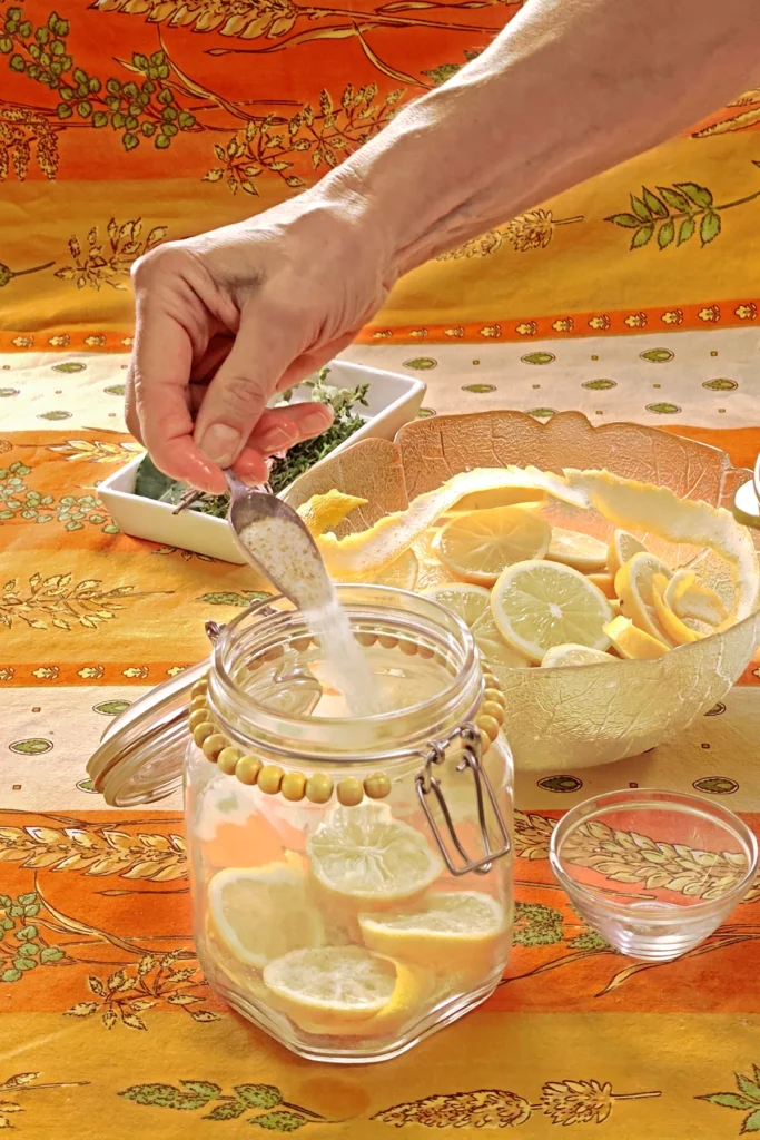 A hand starts filling the preserving jar with the salt and sugar mixture from a spoon. Next to it is a small bowl that will be used as a weight to press the lemon slices together in the end. Behind the jar is a glass bowl with lemon slices and white bowl with herbs - all set on an orange tablecloth with wheat and oat straw motifs.