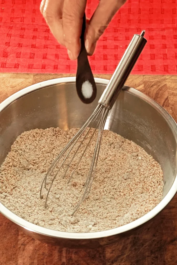 A hand holds a wooden spoon with salt and adds it to a stainless steel bowl with a whisk. The background is a red tablecloth.