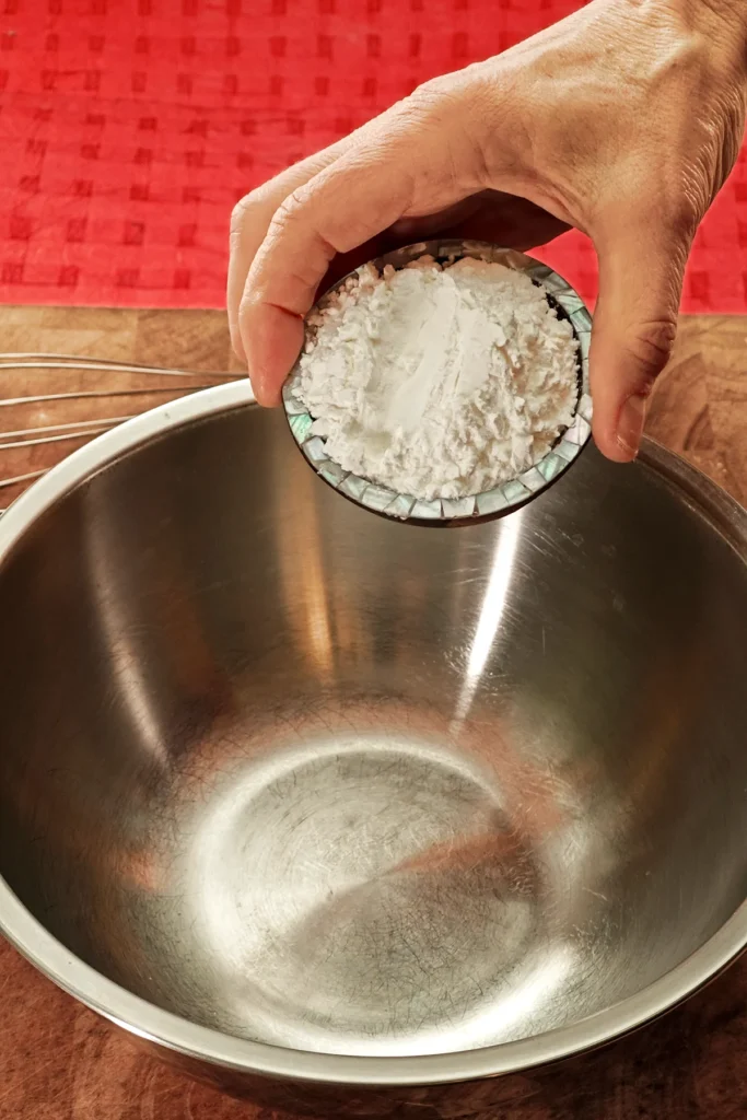 A hand holds a bowl with corn to a stainless steel bowl. The background is a red tablecloth.