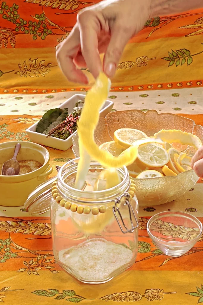 A hand starts filling the preserving jar with a long lemon peel. Next to it is a small bowl that will be used as a weight to press the lemon slices together in the end. Behind the jar is a glass bowl with lemon slices, a white bowl with herbs, and a yellow bowl with the salt and sugar mixture, all set on an orange tablecloth with wheat and oat straw motifs.