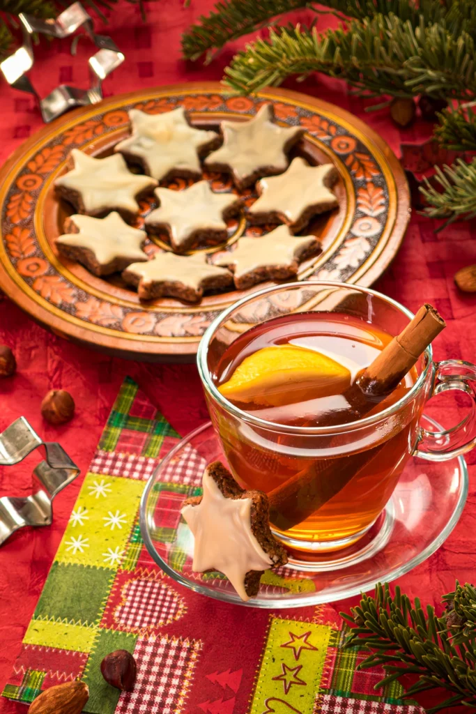 Festively arranged Christmas teatime, with a red tablecloth, fir branches and metal star cookie cutters. In the foreground is a glass teacup with lemon and a cinnamon stick in the tea. On the glass saucer is a cinnamon star leaning against the teacup. Next to it is a paper napkin with a Christmas pattern. Behind it is a wooden plate with more cinnamon stars. Some almonds and hazelnuts are scattered all around.