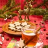 Festively arranged Christmas teatime, with a red tablecloth, a candle, fir branches and decorative elements. In the foreground is a glass teacup with lemon and a cinnamon stick in the tea. On the glass saucer is a cinnamon star leaning against the teacup. Next to it is a paper napkin with a Christmas pattern. Behind it is a wooden plate with more cinnamon stars. Almonds and hazelnuts are scattered all around. Three metal star cookie cutters are scattered around the plate.