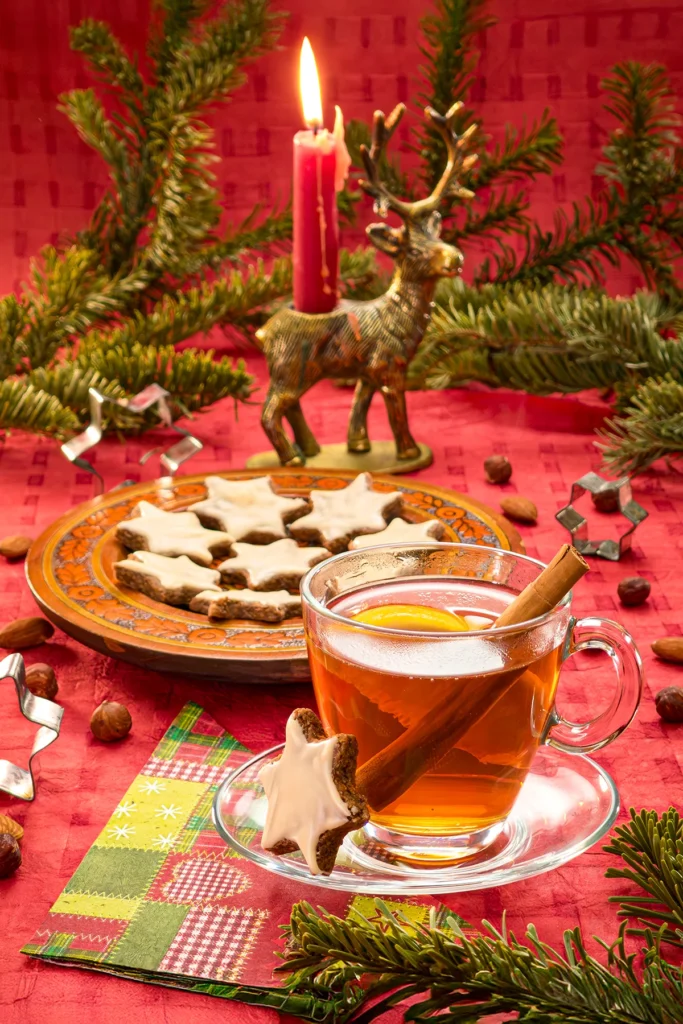 Festively arranged Christmas teatime, with a red tablecloth, a candle, fir branches and decorative elements. In the foreground is a glass teacup with lemon and a cinnamon stick in the tea. On the glass saucer is a cinnamon star leaning against the teacup. Next to it is a paper napkin with a Christmas pattern. Behind it is a wooden plate with more cinnamon stars. Almonds and hazelnuts are scattered all around. Three metal star cookie cutters are scattered around the plate.