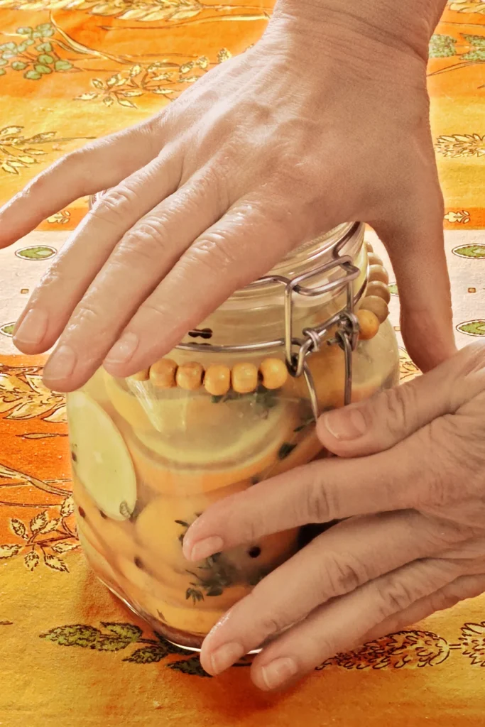 Close-up of a closed preserving jar with lemon slices and herbs. The jar is standing on an orange and yellow patterned tablecloth. Two hands are in the process of closing the swing stopper on the lid.