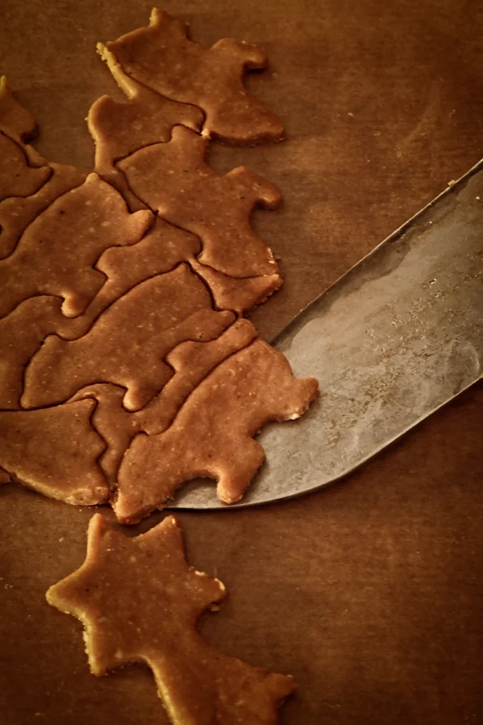 The gingerbread dough is rolled out and placed on baking paper. Many cookies have been cut into the dough, you can see bears and shooting stars. The tip of a large knife removes the cut-out shapes from the dough.