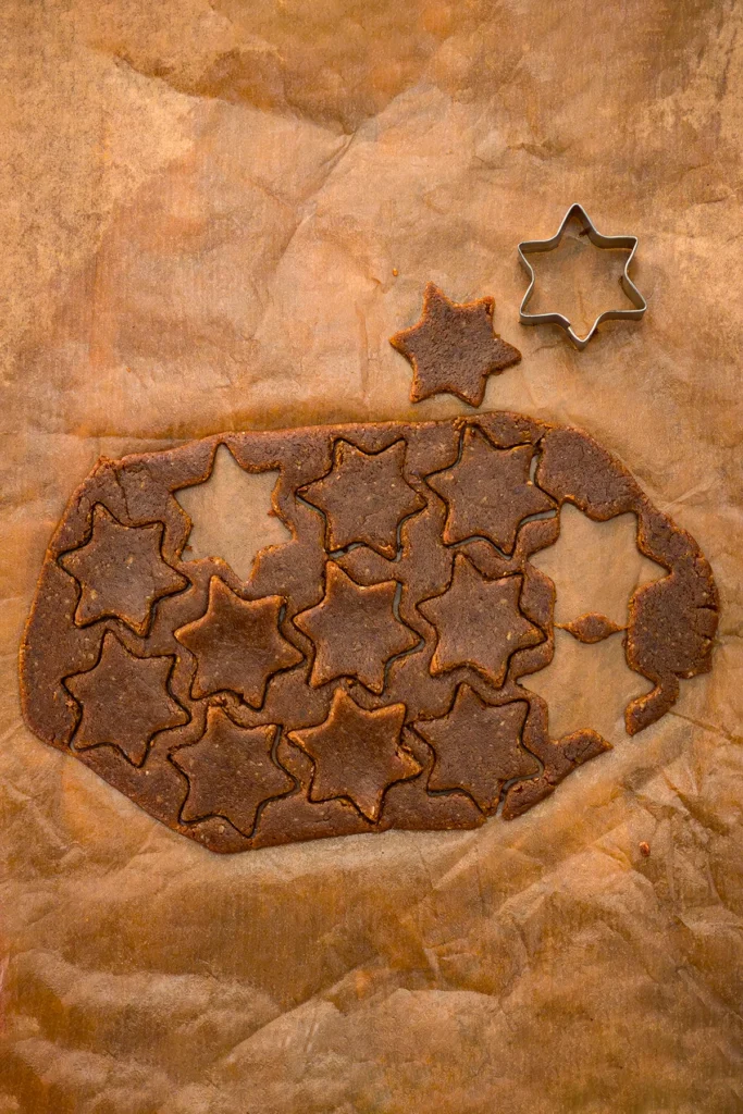 Rolled-out cinnamon star cookie dough with freshly cut star shapes on parchment paper, ready for baking. The star cookie cutter lies next to a cookie.