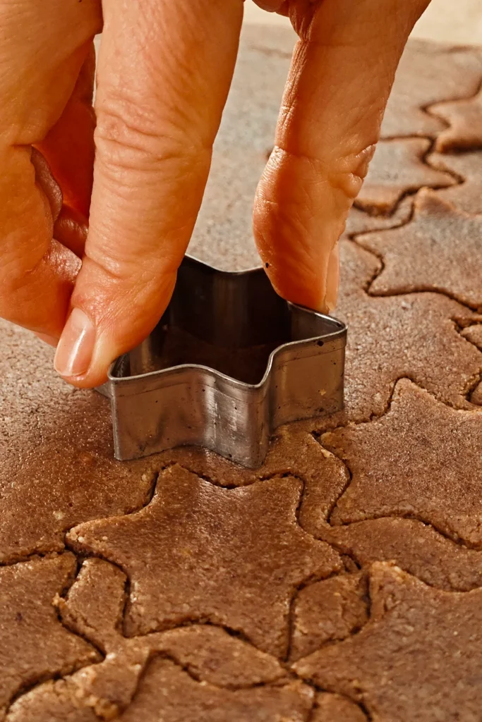 Close-up of hands using a metal star-shaped cookie cutter to cut cinnamon star cookies from rolled-out dough.