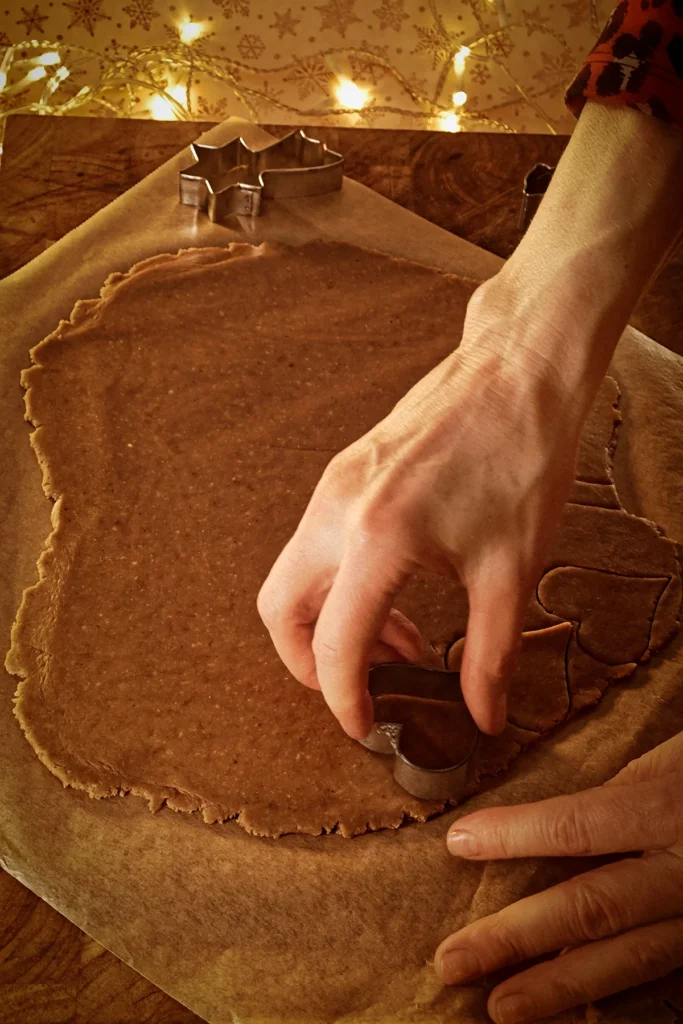 The gingerbread dough is rolled out and placed on baking paper. One hand holds a heart-shaped cookie cutter and cuts hearts into the dough. There are two other cookie cutters in the background.