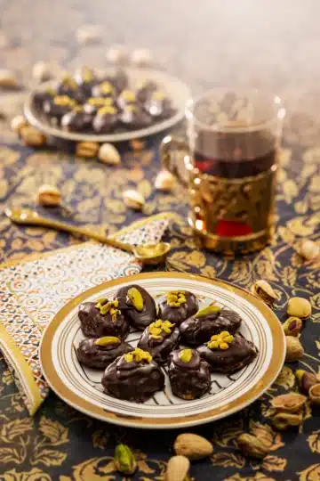 A plate of homemade Dubai-style chocolate truffles, coated in dark chocolate and topped with pistachio halves and chopped pistachios. The truffles are served on a golden striped plate, placed on a rich, gold-patterned tablecloth. In the background, a glass of tea in a golden holder and additional truffles on a separate plate add warmth and charm to the setting. Scattered pistachios and a decorative napkin complete the inviting scene.