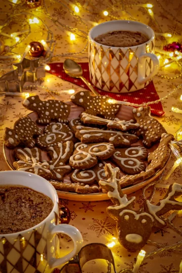 Festive gingerbread cookies decorated with vegan icing, served on a holiday-themed plate alongside two cups of hot cocoa, surrounded by warm fairy lights and Christmas ornaments.