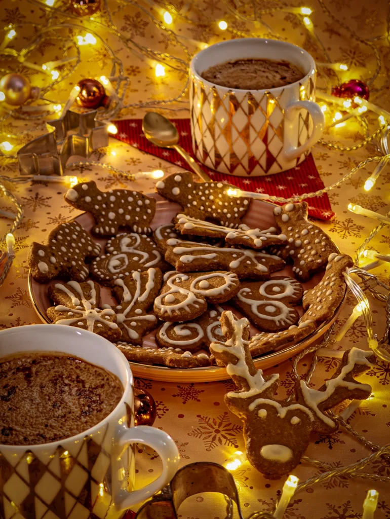 Festive gingerbread cookies decorated with vegan icing, served on a holiday-themed plate alongside two cups of hot cocoa, surrounded by warm fairy lights and Christmas ornaments.