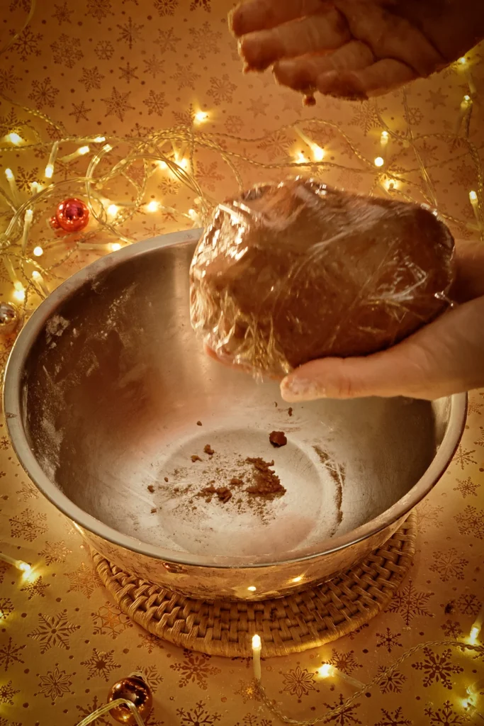 Two hands wrap the gingerbread dough in plastic foil over a stainless steel bowl. A string of lights shines in the background and a few small Christmas baubles are scattered around. The light-colored tablecloth has a festive golden star motif.